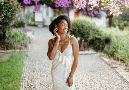 Téléchargez les photos : Heureuse femme afro-américaine millénaire en robe se promène dans le parc en fleurs, bénéficie de voyages d'été et de temps libre le week-end, en plein air. Voyage seul, mode de vie et soins de beauté, annonce, offre - en image libre de droit