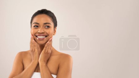 Foto de Rutina de cuidado. Sonriente hermosa mujer negra joven envuelta en toalla de baño blanca tocando su cara y sonriendo, mirando el espacio de copia, aislado en el fondo gris del estudio, banner web - Imagen libre de derechos