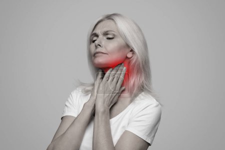 Kranke reife Dame mit Halsschmerzen, Berührung ihres Halses mit rot entzündeter Zone, die an Kehlkopfentzündung, Mandelentzündung, Kehlkopfkrebs, Erkältung leidet, posiert auf Studiohintergrund, Schwarz-Weiß-Foto