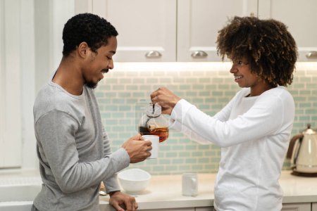 Téléchargez les photos : Beau couple millénaire afro-américain bel homme et jolie femme en pyjama dégustant du thé aromatique ensemble à la maison, intérieur de cuisine confortable. Amour, famille, relations - en image libre de droit