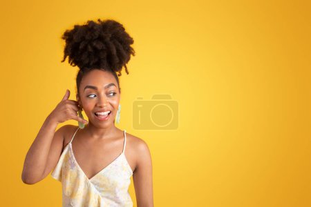 Foto de Alegre dama afroamericana milenaria en vestido haciéndome llamarme gesto, coqueteando, aislado sobre fondo amarillo, estudio. Buenas noticias, chismes, señales de comunicación, anuncios y ofertas - Imagen libre de derechos