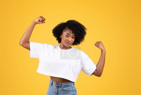 Photo for Cheerful strong young black lady in casual show muscles on arms, biceps, isolated on orange studio background. Power, sport, human emotions, active lifestyle, ad and offer - Royalty Free Image