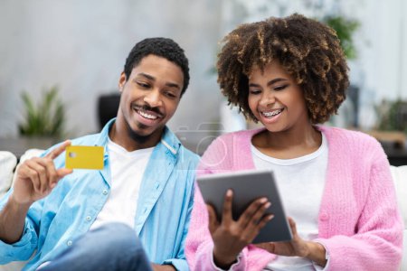 Photo for Cheerful beautiful african american lovers young man and woman sitting on couch at home, using digital tablet, holding yellow plastic bank card. Internet banking concept - Royalty Free Image