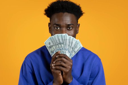 Photo for Portrait of young african american man wearing casual blue sweater showing bunch of cash dollar banknotes. Black guy got cashback, won money, isolated on yellow studio background - Royalty Free Image