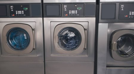 Foto de Servicio de lavandería pública. Fondo con tres lavadoras industriales en la sala de lavandería de pie en una fila en el interior. Concepto de equipo y tecnología de lavandería. Panorama - Imagen libre de derechos
