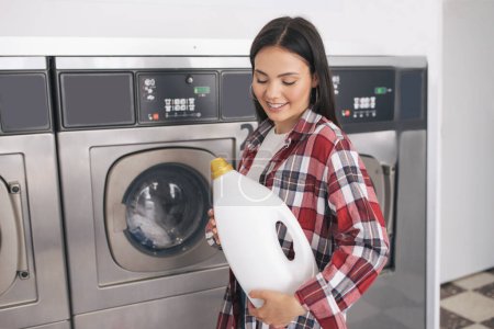 Photo for Laundromat. Young Woman Smiling Holding Detergent Bottle, Cleaning And Washing Clothes At Public Laundrette Service, Standing At Row Of Industrial Washer Machines Indoors - Royalty Free Image