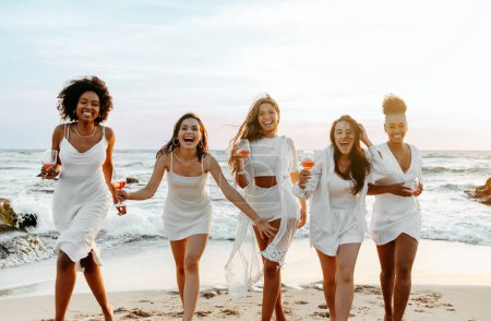 Téléchargez les photos : L'équipe des mariées. Groupe de dames heureuses dansant et s'amusant sur la plage, faisant la fête des poules sur le littoral. Des amies profitent de leurs vacances au bord de l'océan - en image libre de droit