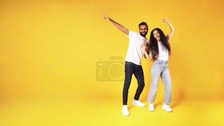Photo for Cheerful middle eastern young couple posing and pointing finger aside at copy space on yellow studio background. Full length shot of joyful millennial man and woman advertising text offer. Panorama - Royalty Free Image