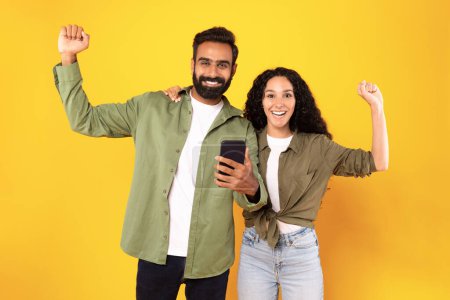 Téléchargez les photos : Heureux jeune couple arabe célébrant victoire tenant smarphone et secouant les poings serrés sur fond jaune, plan studio. Joyeux époux lisant un message au téléphone et faisant un geste oui - en image libre de droit