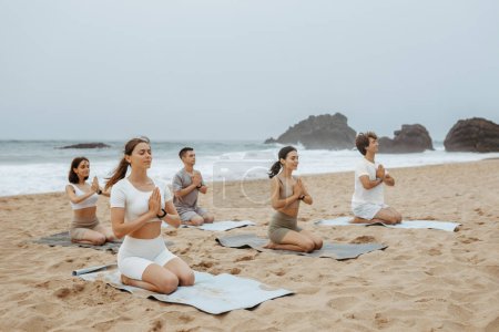 Foto de Yoga en grupo sentado y meditando en la playa, practicando asana con las manos unidas, personas disfrutando del entrenamiento matutino en la costa. Concepto de concentración y serenidad - Imagen libre de derechos