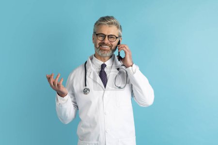 Photo for Telemedicine and Virtual Consultations. Friendly handsome middle aged grey-haired doctor have phone call with patient, consulting remotely, smiling and gesturing over blue studio background - Royalty Free Image