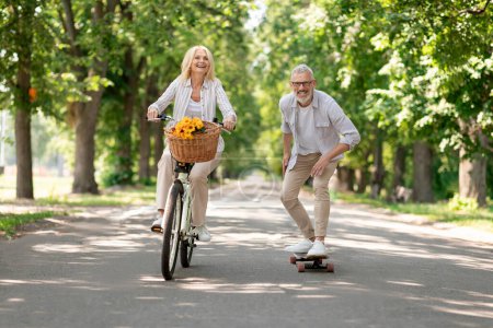 Pensionistas modernos. Bicicleta de montar en pareja feliz y monopatín en el parque, hombre maduro alegre y mujer que se divierten al aire libre juntos, disfrutando de un estilo de vida activo en la jubilación, Copiar el espacio