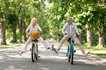 Active Retirement. Happy senior couple having fun while riding bicycles at summer park, cheerful mature man and woman cycling on retro bikes and laughing, enjoying outdoor leisure, copy space