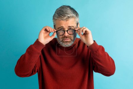 Téléchargez les photos : Homme d'âge moyen tient ses lunettes pour vérifier parce qu'il ne règle pas la mise au point avec sa vue sur le bleu. Problème de vue. Optométrie. Lunettes avec lentilles convexes ou concaves. Myopie. Maladie oculaire. - en image libre de droit