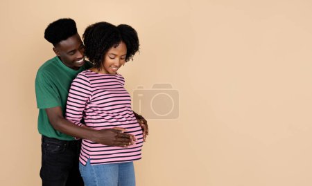 Foto de Feliz millennial afroamericano marido abrazando señora con gran vientre, sintiéndose bebé, aislado sobre fondo beige, estudio. Cuidado de la salud, familia, tratamiento de la infertilidad, embarazada - Imagen libre de derechos