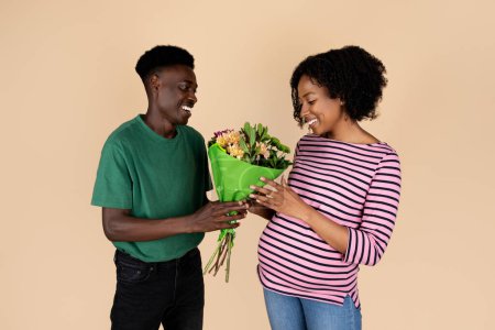 Photo for Glad millennial african american man gives bouquet of flowers to wife with big belly, isolated on beige background. Romantic relationship, date, celebration anniversary, birthday, expect child - Royalty Free Image
