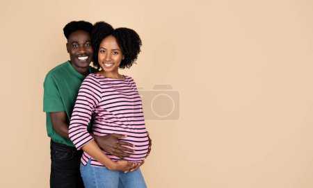Foto de Feliz millennial afroamericano marido abrazando mujer con gran vientre, aislado sobre fondo beige, estudio. Relaciones familiares, buenas noticias, tratamiento de infertilidad, esperando un bebé - Imagen libre de derechos