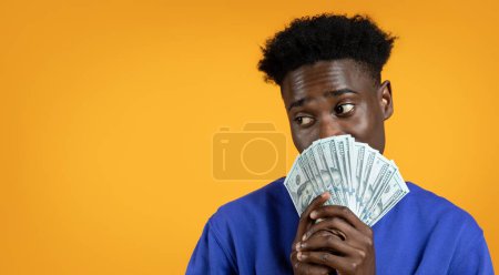 Photo for Lottery Luck. Happy young black man has just won the lottery jackpot, holding bunch of cash dollar banknotes over his face, looking at copy space, isolated on yellow studio background, banner - Royalty Free Image
