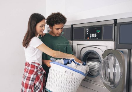 Photo for Laundromat Offer. Young Man And Woman Folding Laundry Doing Chores Together At Laundrette Indoor. Couple With Basket Loading Washing Machine Putting Clothes Inside Washer On Weekend - Royalty Free Image