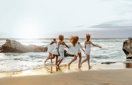 Photo for Happy young female friends having fun and celebrating hen party while running on the beach at the sunset, enjoying evening at coastline, free space - Royalty Free Image