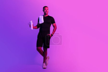 Téléchargez les photos : Pleine longueur de beau sportif noir avec bouteille d'eau souriant posant avec serviette sur fond de studio violet. Garçon en forme positive dans fitwear et boisson de sport debout près de l'espace de copie - en image libre de droit