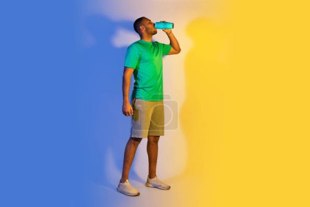 Photo for Sport, Hydration And Weight Loss. Full Length Of Young Black Sportsman Drinking Water From Bottle, Hydrating During Workout Training Standing Over Blue And Yellow Studio Background, Free Space - Royalty Free Image