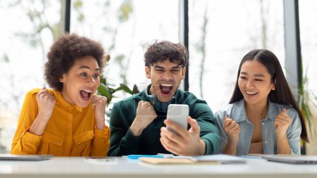 Foto de Tres estudiantes multirraciales muy alegres revisando los resultados de los exámenes en línea en el teléfono inteligente, obteniendo excelentes notas y apretando los puños, sentado en la mesa en el aula universitaria, panorama - Imagen libre de derechos