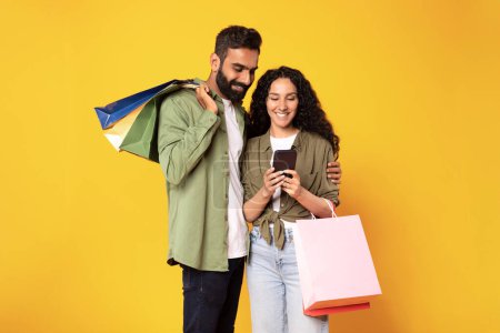 Téléchargez les photos : Mobile Shopping App. Couple arabe heureux utilisant un téléphone portable achetant quelque chose en ligne dans l'application debout avec des sacs à provisions colorés sur fond jaune, Studio Shot. Commerce électronique - en image libre de droit