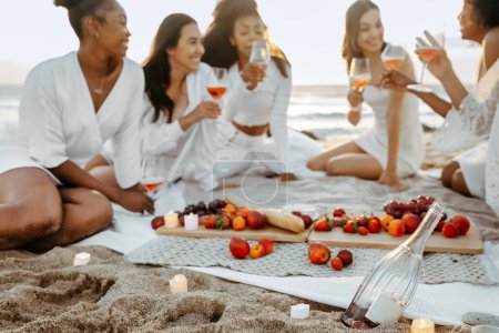 Téléchargez les photos : Groupe de joyeux yougn femmes appréciant manger des fruits et boire du vin rose sur la plage pique-nique d'été, ayant fête bachelorette - en image libre de droit