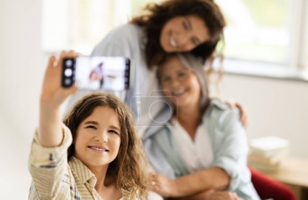 Photo for Smiling senior, millennial women and teenage girl hugs, make selfie on phone screen in living room interior. Family, love, female generation, relationship, video call app and social networks - Royalty Free Image