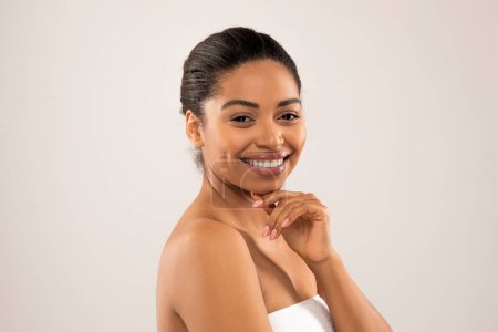 Téléchargez les photos : Joyeux attrayant afro-américain jeune femme enveloppée dans une serviette de bain blanche isolée sur fond de studio gris, toucher son menton et sourire à la caméra, profiter des résultats du traitement spa - en image libre de droit