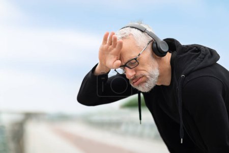 Photo for Photo of tired exhausted sportive pensioner man take break, catch heavy breath, touching his head, elderly sportsman training jogging outdoors, using wireless headphones, copy space - Royalty Free Image