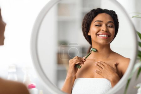 Téléchargez les photos : Belle jeune femme noire massant le cou avec le rouleau de jade de Greenstone à la maison, souriant femme afro-américaine millénaire regardant le miroir et utilisant l'outil de beauté pour les soins de la peau, gros plan - en image libre de droit