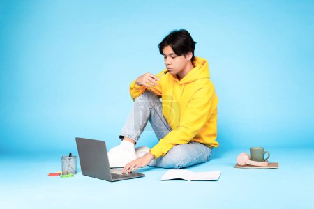Foto de Pensativo chico adolescente coreano usando el ordenador portátil y pensando en sostener la pluma cerca de la boca, sentado en el suelo y haciendo la tarea en línea en el fondo del estudio azul. E-Learning Rutina, Sitio Web Educativo Anuncio - Imagen libre de derechos