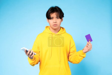 Téléchargez les photos : Acheteur mécontent. Coréen adolescent gars tenant smartphone et carte de crédit exprimant la déception et l'émotion négative, n'ayant pas d'argent sur le compte bancaire sur fond bleu studio. Opération échouée - en image libre de droit