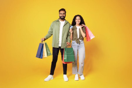 Photo for Shopping. Smiling middle eastern couple with credit card and shopper bags posing, bought a lot of purchases during seasonal sale over yellow background in studio. Full length shot - Royalty Free Image