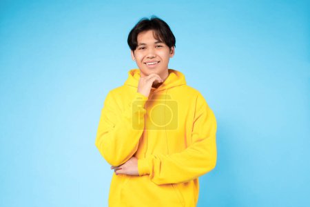 Téléchargez les photos : Portrait d'ado. gai asiatique adolescent guy pose tenant la main près menton regardant caméra avec heureux sourire debout portant jaune sweat à capuche sur bleu fond, studio tir - en image libre de droit