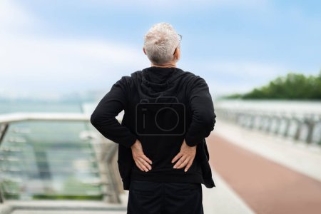 Photo for Exhausted elderly grey-haired sportsman wearing black sportswear touching lower back, senior man exercising outdoor, jogging by footbridge. Exercise and injury prevention in older people - Royalty Free Image