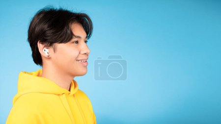 Téléchargez les photos : Korean Teenager Boy Wearing Earbuds Earphones Écoute de la musique, Portrait de vue latérale sur fond bleu. Asiatique adolescent relaxant et profiter favori chanson à la recherche à côté de l'espace libre, panorama - en image libre de droit