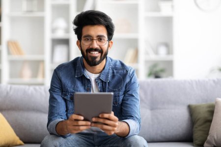 Photo for Handsome Indian Guy Holding Digital Tablet Computer And Smiling At Camera, Sitting On Couch At Home, Using Modern Technologies For Leisure, Browsing Internet Or Checking Social Networks, Free Space - Royalty Free Image