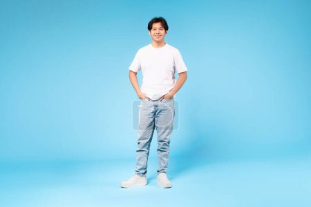 Foto de Tiro de longitud completa de confiado sonriente adolescente japonés de pie posando con las manos en los bolsillos, mirando a la cámara sobre fondo azul en el estudio, usando pantalones vaqueros y camiseta, espacio de copia - Imagen libre de derechos