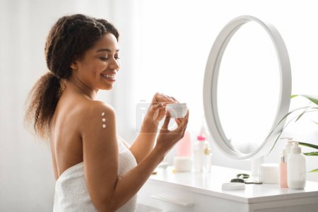 Téléchargez les photos : Nourrissant la peau. Attrayant noir femme appliquant crème hydratante ou lotion sur l'épaule, sourire belle afro-américaine enveloppée dans une serviette après le bain faire des traitements de beauté à la maison - en image libre de droit
