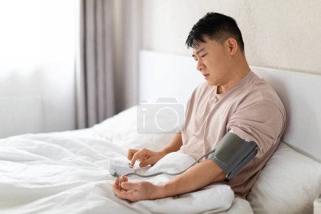 Photo for Unhealthy chinese middle aged man wearing pajamas sitting in bed, checking his blood pressure with modern electronic tonometer. Doing morning checkup after waking up or feeling sick, copy space - Royalty Free Image