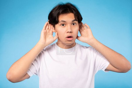 Photo for Huh, What Did You Say. Asian Teen Guy Listening Holding Hands Near Ears Looking At Camera Over Blue Studio Background. Teenager Student Making Dont Hear You Gesture. Overhearing, Speak Up Concept - Royalty Free Image