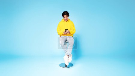Photo for Application Ad. Happy Chinese Teen Guy Using Smartphone Websurfing And Texting Sitting In Studio On Blue Background. Student Using Mobile App And Networking In Social Media Via Phone, Panorama - Royalty Free Image