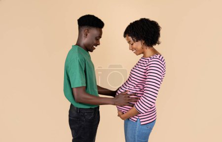 Foto de Positivo millennial afroamericano chico abrazando a la mujer con el vientre grande, se siente niño movimiento, aislado sobre fondo beige, estudio. Recién nacido, relaciones familiares, tratamiento de infertilidad, esperando un bebé - Imagen libre de derechos