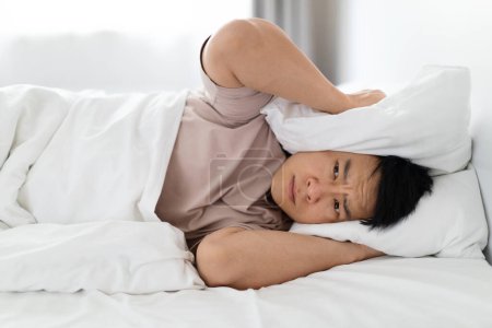 Photo for Angry furious sleepless middle aged asian man wearing pajamas lying in bed at home, covering head with pillow, cannot sleep because of partner snoring or noise from outside, closeup - Royalty Free Image