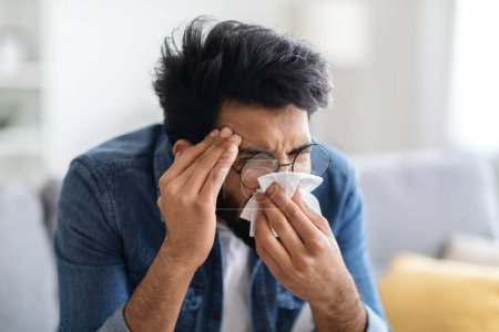 Téléchargez les photos : Grippe saisonnière. Malade Indien soufflant nez liquide dans la serviette tandis qu'il était assis sur le canapé, le jeune homme de l'Est malade se sentant mal à l'aise à la maison, homme millénaire ayant des symptômes de rhume ou de grippe, gros plan - en image libre de droit