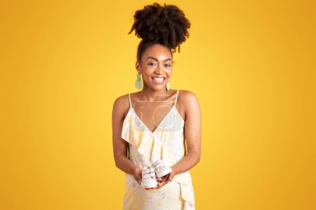 Photo for Smiling pretty millennial african american lady show small shoes, isolated on yellow background, studio. Ad and offer, pregnancy, motherhood, expectation of child and fertility - Royalty Free Image