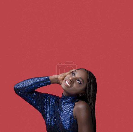 Foto de Retrato de mujer negra joven sonriente soñadora con vestido de fiesta de lentejuelas, atractiva mujer afroamericana con cabello trenzado y maquillaje artístico mirando hacia arriba en el espacio de copia, posando sobre fondo rojo - Imagen libre de derechos